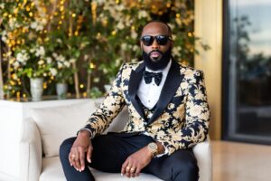 Homme en smoking fleuri, lunettes de soleil, assis avec confiance