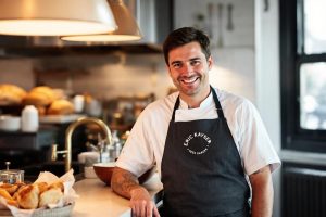 Chef en tablier noir posant dans une cuisine lumineuse